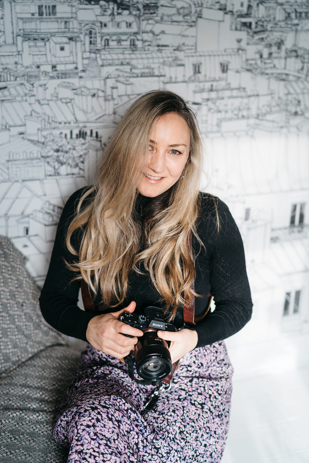 Miss Gen Photography. A woman with long blonde hair holds a black DSLR camera while sitting on a gray textured couch in front of a black and white cityscape line art wall mural. Genevieve the artist behind Miss Gen & KOKO Studio fine art prints.