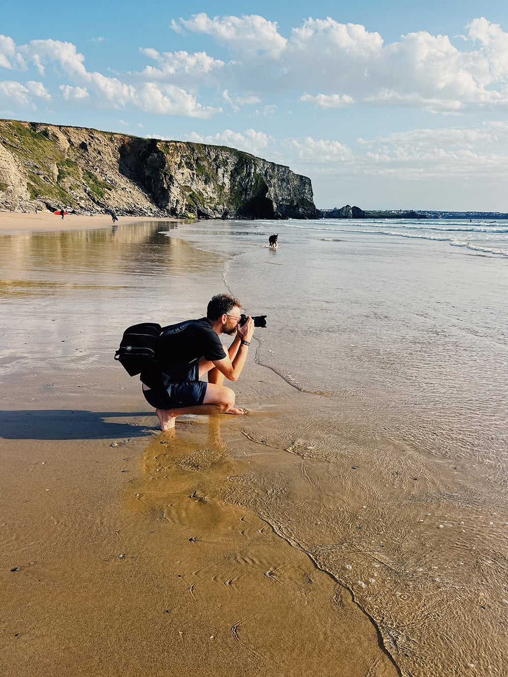 A man crouches in shallow beach water at Watergate Bay in Cornwall, holding a camera, with a rocky coastline, sandy shore, and blue sky with clouds in the background.